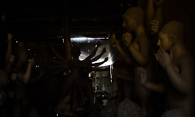 Numerous clay sculptures with human-like features are arranged in a dimly lit workshop. Some figures have multiple arms, suggesting they depict deities or mythological characters. The space is partially illuminated by light filtering through an opening, creating a dramatic play of light and shadow on the sculptures.