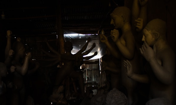 Numerous clay sculptures with human-like features are arranged in a dimly lit workshop. Some figures have multiple arms, suggesting they depict deities or mythological characters. The space is partially illuminated by light filtering through an opening, creating a dramatic play of light and shadow on the sculptures.