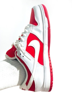 A white and red athletic shoe with a prominent swoosh logo is worn on a foot. The shoe features a lace-up design, primarily white with red accents on the heel, toe, and sole. The fabric appears sleek and clean, with a textured sole for grip.
