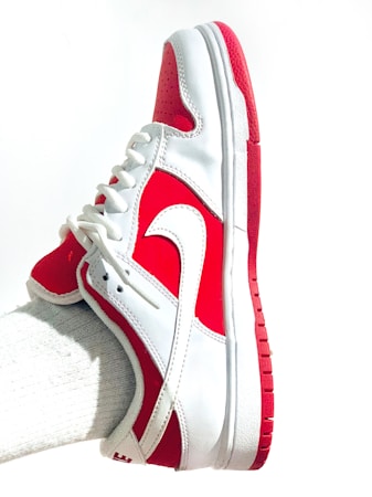 A white and red athletic shoe with a prominent swoosh logo is worn on a foot. The shoe features a lace-up design, primarily white with red accents on the heel, toe, and sole. The fabric appears sleek and clean, with a textured sole for grip.