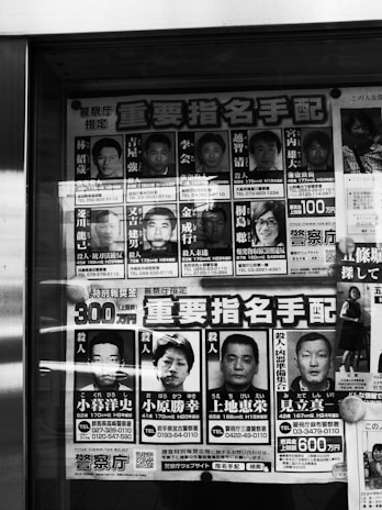 A poster displaying several headshots of individuals accompanied by text, likely in Japanese. The poster is monochrome, featuring both men and a woman, with names and other details printed next to each photo. The photos appear to be mugshots or identification photos.