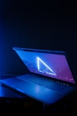 A partly open laptop with a glowing screen displaying a triangular neon light against a dark background. The surrounding area is illuminated by a cool blue hue.