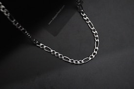 A silver chain with interlocking links is laid out on a dark, almost black background. The lighting creates subtle reflections on the metal surface.