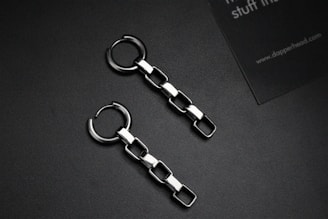 Two metallic chain earrings with square links are placed on a dark surface. Alongside is a partial view of a business card with text.