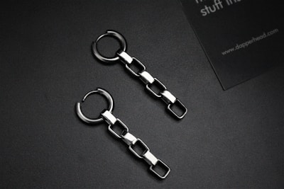 Two metallic chain earrings with square links are placed on a dark surface. Alongside is a partial view of a business card with text.