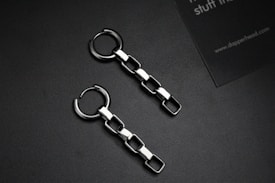 Two metallic chain earrings with square links are placed on a dark surface. Alongside is a partial view of a business card with text.