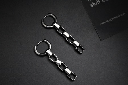 Two metallic chain earrings with square links are placed on a dark surface. Alongside is a partial view of a business card with text.