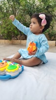 A young child wearing a light blue outfit sits on a white mat outdoors. The child is holding a colorful toy and appears to be reaching forward. A large pink bow is in the child's hair. A lush green background with plants adds vibrancy to the scene.