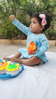 A young child wearing a light blue outfit sits on a white mat outdoors. The child is holding a colorful toy and appears to be reaching forward. A large pink bow is in the child's hair. A lush green background with plants adds vibrancy to the scene.