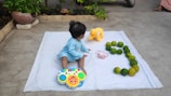 A young child is sitting on a white blanket in an outdoor setting. The child is wearing a light blue outfit and playing with a colorful toy piano. Nearby on the blanket, there is a plush yellow elephant, a pink headband, and a series of green citrus fruits arranged in the shape of the number 3. The background includes a garden area with plants and some concrete pavement.