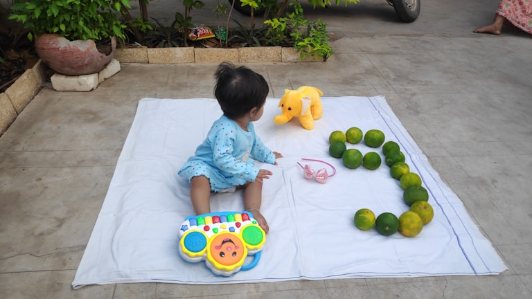 A young child is sitting on a white blanket in an outdoor setting. The child is wearing a light blue outfit and playing with a colorful toy piano. Nearby on the blanket, there is a plush yellow elephant, a pink headband, and a series of green citrus fruits arranged in the shape of the number 3. The background includes a garden area with plants and some concrete pavement.