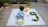 A young child is sitting on a white blanket in an outdoor setting. The child is wearing a light blue outfit and playing with a colorful toy piano. Nearby on the blanket, there is a plush yellow elephant, a pink headband, and a series of green citrus fruits arranged in the shape of the number 3. The background includes a garden area with plants and some concrete pavement.
