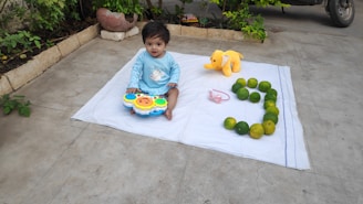 A child is sitting on a white mat on the ground, holding a colorful toy with a face on it. Next to the child is a yellow stuffed elephant, and a number three is formed using green fruits. A pink bow is placed on the mat. The background includes some potted plants and part of a vehicle.