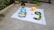 A child is sitting on a white mat on the ground, holding a colorful toy with a face on it. Next to the child is a yellow stuffed elephant, and a number three is formed using green fruits. A pink bow is placed on the mat. The background includes some potted plants and part of a vehicle.
