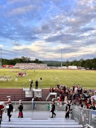 A football field with teams preparing for a game, surrounded by cheerleaders and a marching band dressed in uniforms. Spectators are seated on bleachers, and the sky is partly cloudy with a mix of blue and gray tones. The field is marked clearly with yard lines, and floodlights are present.