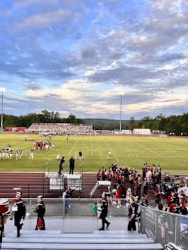 A football field with teams preparing for a game, surrounded by cheerleaders and a marching band dressed in uniforms. Spectators are seated on bleachers, and the sky is partly cloudy with a mix of blue and gray tones. The field is marked clearly with yard lines, and floodlights are present.
