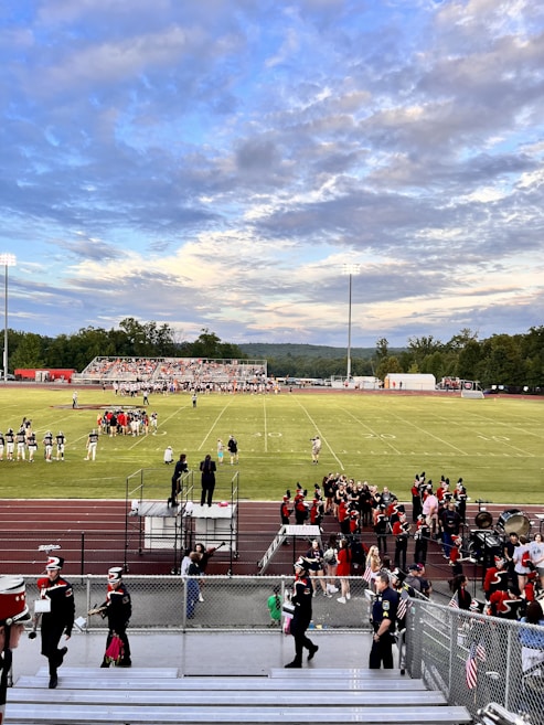 A football field with teams preparing for a game, surrounded by cheerleaders and a marching band dressed in uniforms. Spectators are seated on bleachers, and the sky is partly cloudy with a mix of blue and gray tones. The field is marked clearly with yard lines, and floodlights are present.