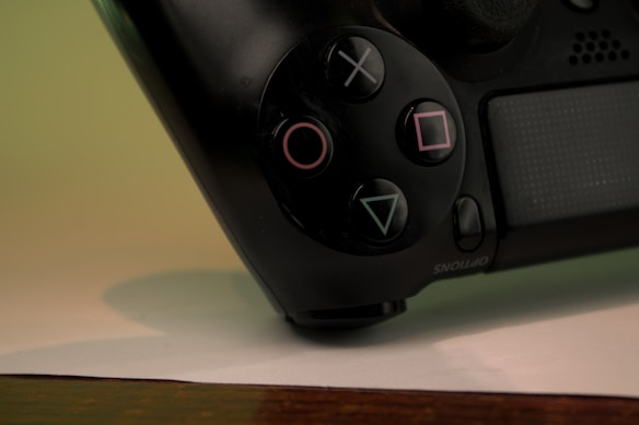 Close-up of a gaming controller showing the iconic PlayStation buttons with triangle, circle, X, and square symbols. The controller is positioned on a flat surface, with a blurred background in shades of green.