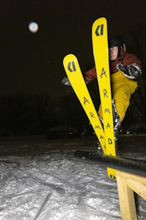 A skier wearing a helmet and goggles is performing a trick on yellow skis with the brand name 'Armada' visible. The scene is set at night, captured in mid-air against a snowy background with some trees visible.
