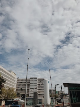A cityscape featuring multiple modern buildings under a partly cloudy sky. The scene includes a tall structure with an 'H' logo and an adjacent cylindrical tower. Various antennas and poles are visible, along with trees and other infrastructure.