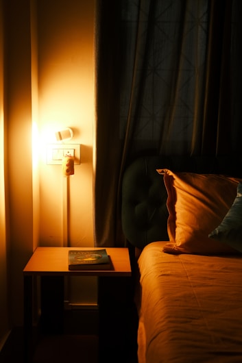 A cozy bedroom corner featuring a nightstand with a book on top, next to a bed with a neatly arranged pillow. The room is warmly lit by a lamp affixed to the wall, casting a gentle glow on the curtains and creating an intimate and calming atmosphere.