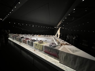 A dimly lit exhibition space features a long, U-shaped table covered with various colored cloths and displays. Each section of the table has decorative items like plates, goblets, and sculptural objects. The space is illuminated by spotlights on the ceiling, casting a focused glow onto the table. Throughout the room, silhouetted figures of onlookers are present, observing the display.