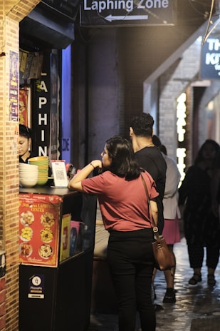 A bustling food stall under the sign 'Laphing Zone' with people waiting for their orders. The atmosphere appears lively, with colorful dishes displayed on a menu. Customers seem engaged and are conversing with the vendor.