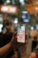 A hand is holding a smartphone with the screen displaying various app icons, including a weather widget indicating a temperature of 34°C and a calendar. The background is blurred, suggesting an urban setting with people and bright lights.