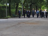 A group of people, including nuns wearing traditional habits, walk together down a tree-lined path. The surrounding area is lush with green foliage, and there are park benches along the sidewalk.