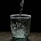 Water is being poured into a clear glass on a dark surface, creating bubbles and splashes inside the glass. The background is completely black, highlighting the transparency and clarity of the glass and water.