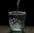 Water is being poured into a clear glass on a dark surface, creating bubbles and splashes inside the glass. The background is completely black, highlighting the transparency and clarity of the glass and water.