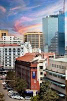 A cityscape featuring a mix of modern and older architecture. The foreground includes a red brick building with a Stanbic Bank sign, surrounded by other commercial structures. A towering glass building dominates the background, reflecting the sky. The sky is vibrant with hues of blue, pink, and orange, indicating a possible sunset. The street below is lined with parked cars and a few pedestrians are visible.