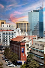 A cityscape featuring a mix of modern and older architecture. The foreground includes a red brick building with a Stanbic Bank sign, surrounded by other commercial structures. A towering glass building dominates the background, reflecting the sky. The sky is vibrant with hues of blue, pink, and orange, indicating a possible sunset. The street below is lined with parked cars and a few pedestrians are visible.