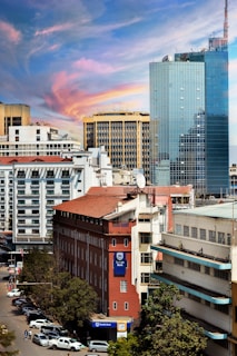A cityscape featuring a mix of modern and older architecture. The foreground includes a red brick building with a Stanbic Bank sign, surrounded by other commercial structures. A towering glass building dominates the background, reflecting the sky. The sky is vibrant with hues of blue, pink, and orange, indicating a possible sunset. The street below is lined with parked cars and a few pedestrians are visible.