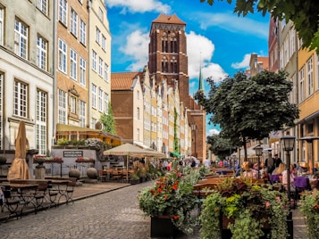 A picturesque European street lined with historic buildings, outdoor cafes, and lush greenery. The cobblestone path is bordered by vibrant flowers. People are enjoying meals and leisurely walking along the street, while a towering brick structure with a spire is visible in the background under a bright blue sky with scattered clouds.