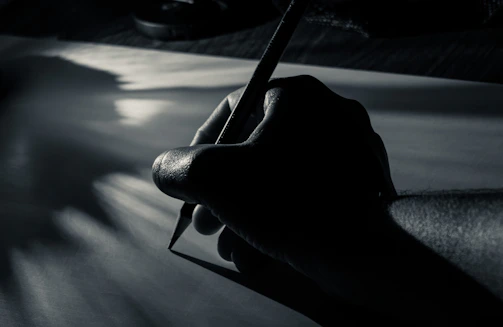 A sleek black and white photo of a hand holding a charcoal pencil poised over paper.