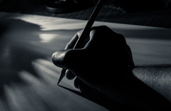 A hand holding a pencil is poised over a blank sheet of paper. The lighting is dramatic, casting deep shadows and highlighting the contours of the hand and pencil. The focus is on the act of writing or drawing, evoking a sense of creativity and concentration.