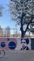 A brick wall features a graffiti mural with a portrait of a person alongside colorful circular designs and text in Spanish. A large tree with green and yellow leaves towers above the wall, casting some shadow. A metal fence stands behind the tree, and part of a bicycle is visible in the bottom left corner.