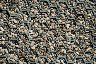 A collection of small, smooth stones arranged within hexagonal metal frames, creating a visually appealing geometric pattern. The stones vary in color from light beige to dark gray, adding a natural variation to the organized structure.