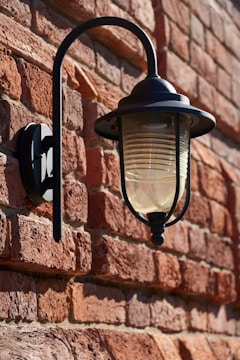 A vintage-style wall-mounted lantern made of metal with a glass enclosure is affixed to a rustic brick wall. The lighting fixture casts a subtle shadow on the textured wall, and natural light highlights the warm red and brown tones of the bricks.