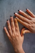 Two hands with decorated fingernails resting on a textured surface. The nails feature a design with black tips and small black dots on a nude base.