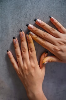 Two hands with decorated fingernails resting on a textured surface. The nails feature a design with black tips and small black dots on a nude base.