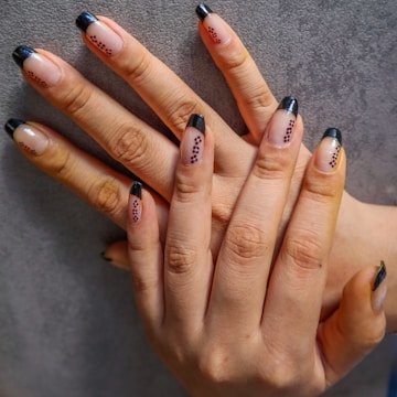 Hands with manicured nails featuring black tips and small decorative black dots on a neutral base. The fingers are interlaced against a textured gray background.
