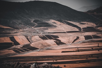 A vast, arid landscape with sweeping fields that are sectioned into irregular plots. The hills have a barren, dusty appearance, and the sky is overcast, casting a muted light across the terrain.