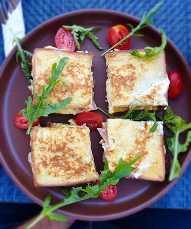 Four slices of grilled sandwiches are arranged on a dark brown plate, garnished with fresh arugula and halved cherry tomatoes. The sandwiches have a golden-brown crispy texture, filled with melted cheese and meat. The plate is set on a textured blue surface, creating a vibrant contrast with the food.