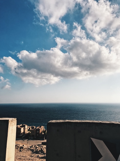 A coastal scene with a vast ocean meeting the horizon under a sky dotted with fluffy clouds. A few residential buildings are visible in the foreground, alongside a concrete barrier.