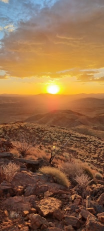 A picturesque landscape featuring a brilliant sunset over rolling hills and rugged terrain. The sky is a mix of vibrant orange and yellow hues with a scattering of clouds. In the foreground, dried grasses and rocky formations are illuminated by the warm glow of the setting sun.
