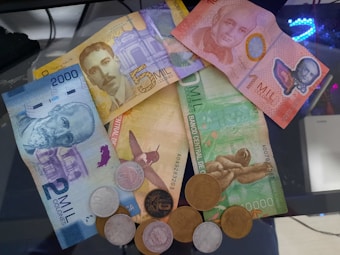 A variety of Costa Rican banknotes and coins are spread on a glass surface. The banknotes are colorful and depict portraits and scenes from nature, such as a hummingbird and a sloth. The coins are mostly silver and bronze in color.