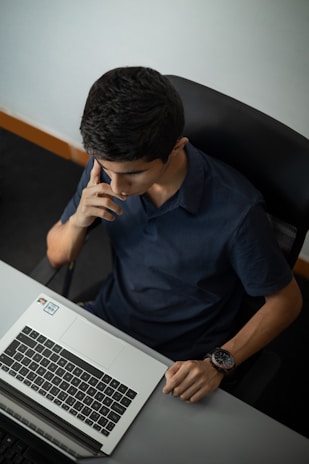 A person seated at a desk is focused on a laptop. They are resting one hand on the desk and appear to be in thought, with their other hand touching their face. The individual is wearing a dark shirt and a wristwatch. The setting looks like an office environment.