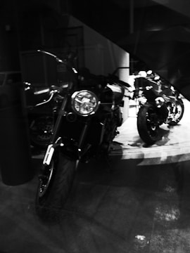 Two motorcycles are parked indoors on a reflective floor, with one prominently in the foreground and partially illuminated. The setting appears dimly lit with limited ambient light.